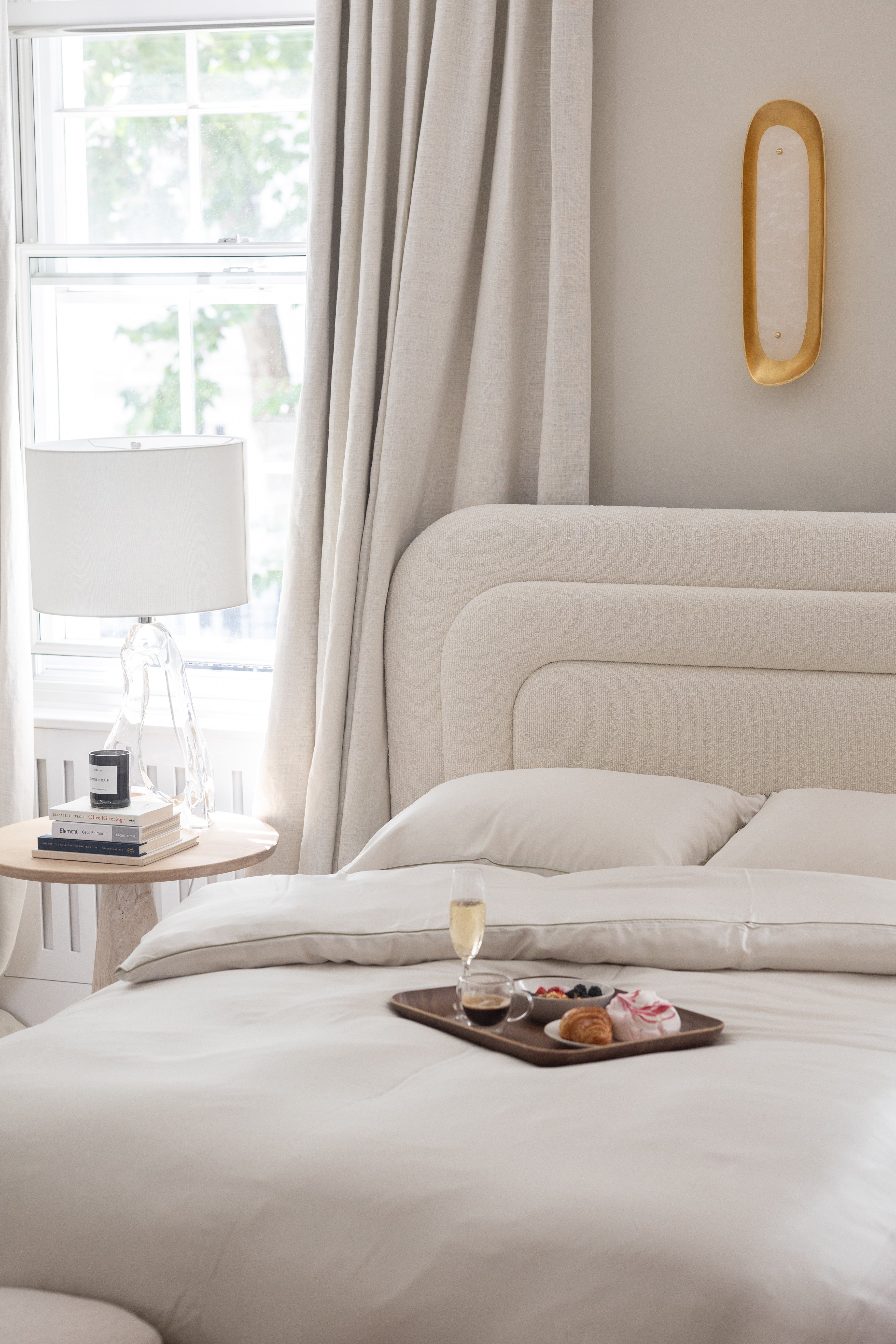Image shows the Dune bed within a bedroom.  The bed features the sculptural curved cornered cream headboard. A side table with a lamp and books is at the left of the bed. A breakfast tray sits on the bed.