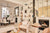 Image shows an open plan living and dining area.  To the left of the image, depicted in  the rear of the space, there are five Kadiev dining chairs finished in off white boucle fabric and antique brass around a Samantha dining table.  The foreground shows a Cecila bench and a Livius armchair both finished in cream shearling fabric, and a Brooke coffee table finished in walnut and travertine. Fireplaces and wall display units featuring various decorative items are in the background. 