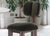 Photograph shows an Elephant Chair with a smoked walnut frame and sage green velvet upholstery in a living area. Large houseplants are in the background. 