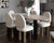 Image shows five Kadiev dining chairs placed around a Samantha dining table.  The chairs’ seat and back cushions are finished in off white boucle fabric and sit on four antique brass legs. A vase with flowers sits on the table.  A sideboard with a lamp is in the background. Artwork is hung on the walls in the background. 